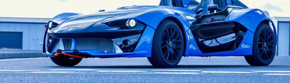 Zenos E10 de couleur bleue vue de 3/4 avant statique dans les stands d'un circuit automobile.